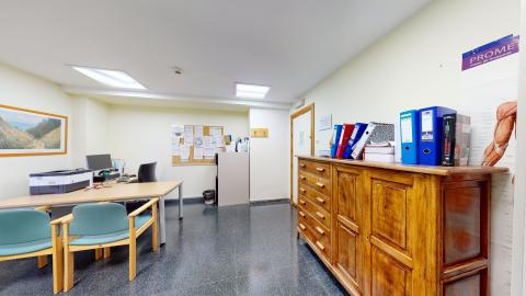 Consulta médica. Consulta médica con paredes de color amarillo pálido y suelo de terrazo oscuro. En el lado izquierdo se sitúa una mesa de despacho con un ordenador, una impresora y dos sillas confidente verdes de madera clara. En la mitad derecha destaca un mueble aparador alto de madera rústica con múltiples cajones y puertas, sobre el que hay archivadores de palanca de colores azul y rojo. En la pared derecha cuelga un póster de anatomía muscular.