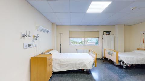 Habitación de enfermería. Una habitación de enfermería amplia con suelo de terrazo oscuro pulido y falso techo con placas blancas e iluminación empotrada brillante. Contiene dos camas articuladas de hospital con barreras de madera clara, dispuestas en paralelo. Las paredes son de color crema, y en la de la izquierda destacan tomas de gases medicinales de pared (oxígeno) y un sistema de llamada de emergencia sobre una mesilla de noche de madera clara. Al fondo, una ventana con persiana veneciana beige deja ver vegetación exterior.