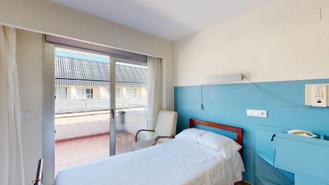 Habitación individual. Una habitación individual muy luminosa con una cama baja vestida de blanco en primer plano. La pared del cabecero, de madera rojiza, tiene un zócalo alto pintado de azul celeste, mientras que el resto de las paredes son de un tono crema claro. A la izquierda, una gran puerta corredera de cristal con cortinas blancas da acceso a una terraza exterior soleada con suelo de baldosas de arcilla y barandilla metálica. Junto a la cama hay un sillón reclinable de color beige con reposabrazos de madera.