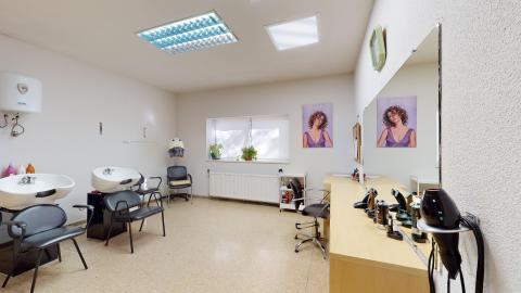 Peluquería. Una sala de peluquería amplia con paredes blancas y suelo de terrazo beige. A la izquierda se sitúan dos lavacabezas con sillones negros tapizados, y al fondo, bajo una ventana luminosa, hay un radiador blanco y plantas decorativas. A la derecha destaca un gran espejo rectangular montado sobre un mueble de madera clara, donde se apoyan secadores de mano, botes de laca y utensilios de peinado. Dos carteles con fotografías de peinados femeninos decoran la pared del fondo.