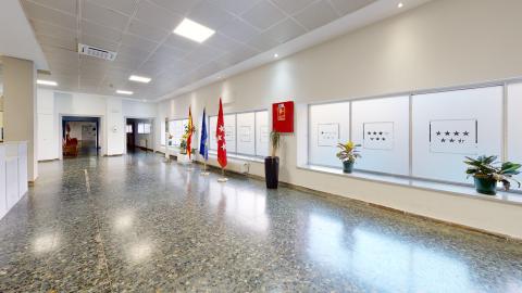 Vestíbulo. Un pasillo muy amplio y luminoso con suelo de terrazo brillante de tonos grises y azulados. La pared derecha cuenta con grandes ventanales de cristal translúcido decorados con el escudo de las siete estrellas de la Comunidad de Madrid; sobre el alféizar hay varias plantas en macetas oscuras. En el centro del pasillo destacan tres mástiles alineados con las banderas de España, la Unión Europea y la Comunidad de Madrid. Al fondo a la izquierda se abren accesos a otras estancias comunes.