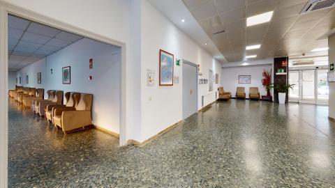Distribuidor y estancia. Un distribuidor interior amplio y bien iluminado que conecta varias salas, con el mismo suelo de terrazo brillante. A la izquierda se abre una gran entrada sin puerta hacia una sala de descanso lineal, donde se alinean numerosos sillones orejeros tapizados en color beige. En las paredes blancas del pasillo cuelgan cuadros decorativos y carteles informativos. Al fondo a la derecha se aprecian la puerta acristalada de acceso exterior y otra pequeña zona de espera con sillones.