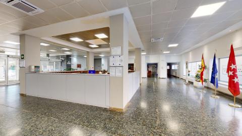 Recepción. Vista amplia de la recepción de la residencia, un espacio muy luminoso con suelo de terrazo oscuro brillante y paredes blancas. A la izquierda destaca un gran mostrador blanco en forma de L, y a la derecha, junto a unos ventanales, se ubican tres mástiles con las banderas de España, la Unión Europea y la Comunidad de Madrid. El techo técnico tiene paneles cuadrados y luces empotradas.