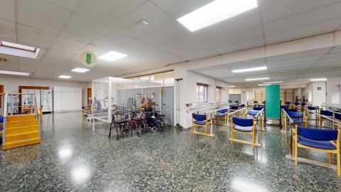Fisioterapia. Amplia sala de fisioterapia muy iluminada, con suelo de terrazo verde oscuro brillante y paredes blancas. El espacio está equipado con diverso material de rehabilitación y fortalecimiento: a la izquierda destaca una rampa con escaleras de madera y una gran jaula metálica blanca con andadores y sillas de ruedas en su base. A la derecha, se suceden varias hileras de paralelas de marcha metálicas con sillas de tapicería azul.
