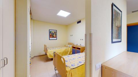 Habitación doble. Interior de una habitación doble amplia y muy luminosa, con paredes de color amarillo pálido y suelos de terrazo claro. Se observan dos camas articuladas individuales con colchas estampadas en amarillo y azul. Entre ambas camas hay dos mesillas de noche de madera clara con cajones azules. De frente dos armarios empotrados a los lados de una ventana con cortinas y, en frente, dos sillas de respaldo alto en azul.