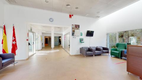 Amplia sala de recepción de paredes blancas con gran luminosidad cenital. A la izquierda destacan las banderas de España y de la Comunidad de Madrid junto a un sillón gris. En el centro, un pasillo con suelo beige se extiende hacia el fondo del edificio. A la derecha de la estancia se ubican tres sillones grises alineados, un televisor apagado en la pared, un sillón de color verde oscuro y el lateral del mostrador de madera.