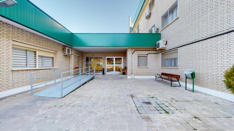 Fachada exterior pavimentada con baldosas de piedra gris bajo un cielo despejado. El edificio es de ladrillo visto de color beige claro y destaca una amplia marquesina metálica de un color verde intenso que corona el porche. A la izquierda, una rampa de accesibilidad metálica de color gris claro con barandillas conduce a la puerta de entrada acristalada. En el lado derecho se sitúan un banco de madera con estructura de hierro negro y una papelera verde.