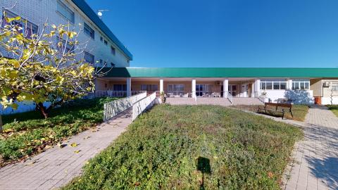 Amplia zona ajardinada de la residencia bajo un cielo azul intenso y soleado. En primer plano hay una pradera de césped verde con un árbol de hojas caducas amarillentas a la izquierda. Un camino de adoquines claros rodea el césped y conecta con una larga rampa peatonal blanca equipada con barandillas. Al fondo, la fachada posterior combina ladrillo beige con ventanales superiores y una planta baja porticada, rematada por la misma marquesina longitudinal verde.