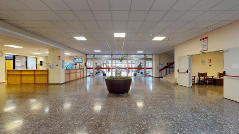 Recepción. Amplio distribuidor interior iluminado por paneles de luz led cuadrados en un techo técnico blanco. El suelo es de terrazo oscuro brillante y en el centro de la estancia destaca un gran macetero circular marrón con plantas verdes. A la izquierda se ubican los mostradores de recepción de madera clara, al fondo la gran cristalera de salida y a la derecha el acceso a unas escaleras interiores.