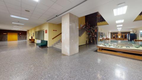 Gran vestíbulo interior muy espacioso y bien iluminado, con suelo de terrazo moteado en tonos grises. En el centro destaca una robusta columna rectangular revestida de placas beige. Las paredes combinan el color amarillo claro con zonas de ladrillo visto oscuro al fondo. Se aprecian unas escaleras de acceso a la planta superior y, en primer término a la derecha, una gran vitrina de madera que exhibe una maqueta del edificio.
