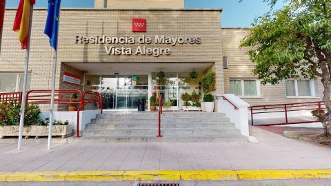 Fachada principal exterior construida con ladrillo visto de tono beige claro bajo una iluminación de día despejado. En la parte superior destacan las letras en relieve «Residencia de Mayores Vista Alegre» junto al logotipo rojo de la Comunidad de Madrid. El acceso cuenta con una escalinata central de piedra gris flanqueada por rampas de accesibilidad con barandillas rojas. La entrada acristalada está decorada con numerosas plantas y macetas.