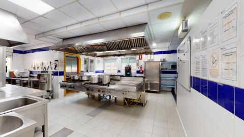 Cocina. Cocina industrial espaciosa y con iluminación blanca directa. Predominan las superficies de acero inoxidable en las encimeras y en la gran isla central con fogones y ollas. Las paredes son de azulejos blancos con una franja horizontal azul oscuro, donde destacan varios carteles informativos y de normativas de seguridad sanitaria.