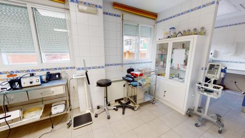 Consulta de enfermería. Consulta de enfermería muy luminosa con dos grandes ventanas equipadas con persianas. El espacio cuenta con paredes de azulejos blancos con cenefa azul y suelo gris claro. Destaca una gran vitrina blanca central llena de material sanitario, flanqueada por una báscula médica de columna a la izquierda y un monitor de signos vitales con ruedas a la derecha.
