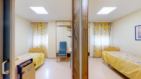 Habitación doble. Habitación doble dividida por un panel central de madera. Es un espacio luminoso con paredes beige y dos ventanas al fondo con cortinas estampadas de flores azules y amarillas. A cada lado del panel destaca una cama articulada individual con colcha amarilla y azul, orientadas de forma simétrica.