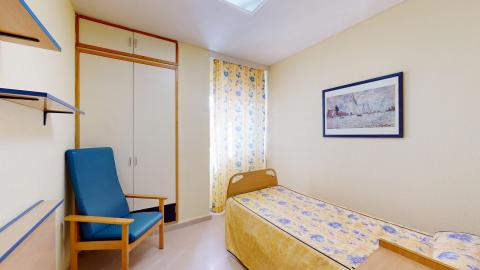 Habitación individual. Habitación individual acogedora y bien iluminada por una ventana con cortina de motivos florales azules y amarillos. Las paredes son de tono beige claro. Destaca una cama articulada con colcha amarilla y un sillón ergonómico tapizado en azul junto a un armario empotrado blanco con perfiles de madera.