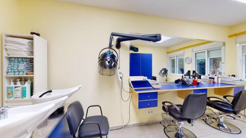 Peluquería. Sala de peluquería luminosa de paredes amarillo pastel y suelo gris. Cuenta con un gran espejo en la pared frontal que refleja el mobiliario azul eléctrico. Destacan dos sillones de peluquería negros con base cromada frente al tocador, un secador de casco de pared y dos lavacabezas blancos en primer plano.