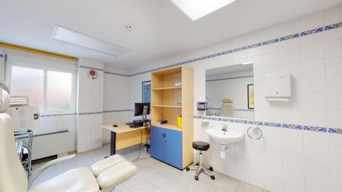 Consulta de podología. Consulta de podología muy iluminada, con paredes de azulejos blancos con una fina cenefa azul y un tragaluz en el techo. Dispone de una ventana de cristal opaco a la izquierda. Destaca en primer plano un sillón clínico de podología blanco y articulado, orientado hacia un escritorio de madera y un mueble bajo de color azul.