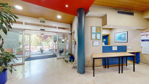 Recepción. Una toma de ángulo amplio de una recepción iluminada. Incluye suelos de baldosas blancas, un mostrador de recepción azul con un escritorio de madera, y una gran puerta de cristal que conduce al exterior con plantas de interior y exterior. Un póster en la pared detrás del mostrador.