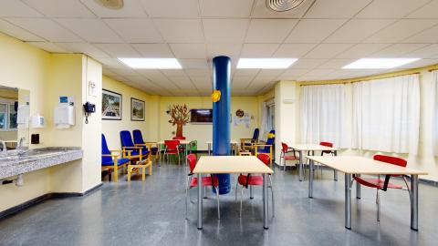 Sala de actividades. Sala de actividades bien iluminada. Tiene paredes de color amarillo claro, suelos grises, mesas y sillas rojas y azules, un fregadero con espejo y una columna azul brillante con una flor naranja. El fondo tiene ventanas con cortinas blancas.