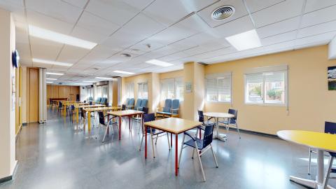 Sala de estar. Sala de estar iluminada. Incluye paredes de color beige claro, un suelo de baldosas gris oscuro, múltiples mesas y sillas de metal con asientos azules, y varias ventanas grandes con persianas que dejan entrar la luz solar.