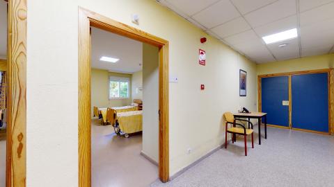 Enfermería. Consulta de enfermería con paredes de color amarillo pálido, suelos grises y beige, un pasillo, una habitación con camas y una ventana, un escritorio de madera con una silla amarilla, y un póster rojo en la pared con un extintor de incendios.