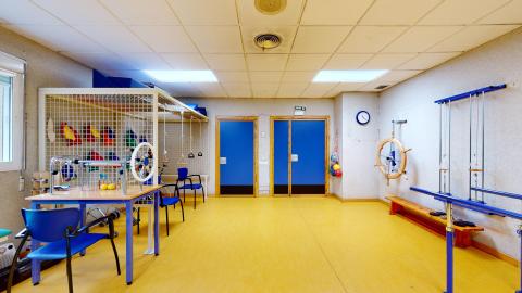 Fisioterapia. Sala de fisioterapia con paredes de color beige, suelos amarillos, dos grandes puertas azules, ventanas con persianas que dejan entrar la luz solar, una variedad de equipos de fisioterapia y un reloj de pared.