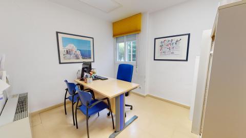 Consulta de psicología. Consulta con paredes blancas, suelos de color beige claro, un escritorio de madera con una silla azul, otra silla azul frente al escritorio, un monitor de computadora, y dos cuadros enmarcados en la pared.