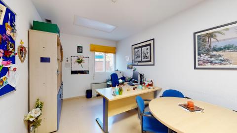 Trabajo social. Sala con paredes blancas, suelos de color beige claro, un escritorio de madera con una silla azul y un monitor de computadora, una gran estantería de madera y azul con varios objetos, y otra mesa redonda de madera con dos sillas azules.