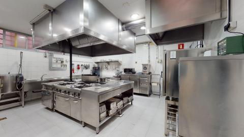 Cocina. Cocina industrial amplia y muy iluminada con luz blanca artificial. Todas las superficies, campanas extractoras, mesas de trabajo y electrodomésticos son de acero inoxidable reluciente. Las paredes están revestidas de azulejos blancos y el suelo es de baldosas gris claro. Destaca la perfecta organización y limpieza de los fogones y zonas de preparación.