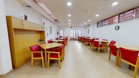 Comedor. Amplio comedor de diseño abierto y muy luminoso gracias a una hilera de ventanales al fondo que dan al jardín. Las paredes combinan pintura blanca con un zócalo de madera clara. El espacio está distribuido con múltiples mesas cuadradas rodeadas por sillas con estructura de madera y tapicería roja. El suelo es de vinilo continuo en tono marrón claro.
