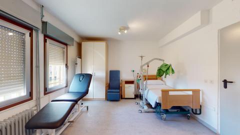 Habitación de enfermería. Habitación clínica individual, espaciosa y bien iluminada por un ventanal al fondo. Consta de una cama hospitalaria articulada con cabecero de madera y una colcha azul. Las paredes están pintadas en un tono verde oliva muy suave y el suelo es de tarima clara. Destaca un sillón de descanso reclinable con tapicería azul junto a la cama y un armario de madera.