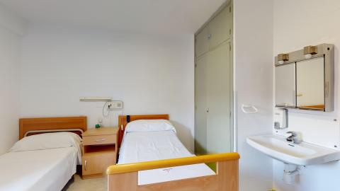 Habitación doble. Dormitorio con dos camas individuales con ropa de cama blanca. Las cabeceras, pieceras y mesilla de noche que las separa son de madera clara. El espacio está pintado en blanco. A la derecha hay un armario empotrado en verde grisáceo claro y a su lado un lavabo con espejo.