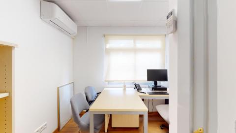 Consulta de psicología. Despacho profesional luminoso con paredes de color beige y suelo de tarima flotante. Está amueblado con un escritorio de madera en ángulo, una silla de oficina ergonómica y un ordenador. Destaca una zona de atención confidencial a la izquierda con dos sillones tapizados en tela azul oscuro y una pequeña mesa auxiliar redonda bajo un ventanal.