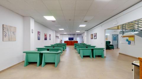 Sala de estar. Amplia sala de estar rectangular iluminada con paneles de luz blanca en el falso techo. Las paredes son blancas y están decoradas con cuadros de flores. El espacio está organizado con numerosas butacas individuales de color verde turquesa dispuestas en filas. Al fondo se observa un mueble de madera con un televisor.
