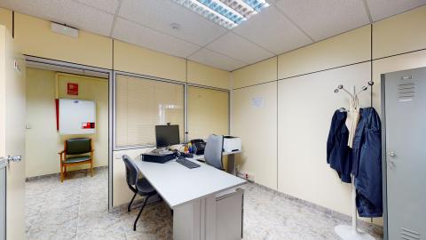 Trabajo social. Despacho de trabajo iluminado con un panel de luz fluorescente. Tiene paredes de paneles de color beige claro y suelo de terrazo gris. Dispone de una mesa de oficina gris con un ordenador, teclado y una silla de escritorio. Destaca un perchero de pie con chaquetas y una taquilla metálica gris a la derecha.