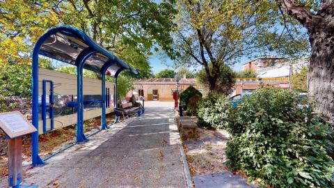 Zona de ejercicio. Zona ajardinada exterior muy soleada y rodeada de árboles frondosos. Un camino de baldosas está flanqueado por vegetación y bancos de piedra. Destaca en primer plano un circuito azul de aparatos de gimnasio al aire libre para mayores, cubierto por una pequeña marquesina.