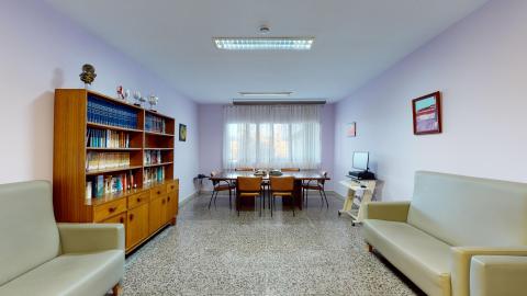 Biblioteca. Sala de lectura amplia con paredes de tono lila claro y suelo de terrazo gris. A la izquierda destaca una gran estantería de madera repleta de libros y trofeos. Al fondo, una gran ventana con cortinas blancas aporta mucha luz natural a una mesa larga de reuniones rodeada de sillas de cuero marrón. Incluye dos sofás de color beige claro y una zona con ordenador.
