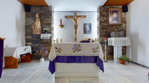 Capilla. Altar de una pequeña capilla con paredes blancas y suelo de mármol claro muy brillante. El altar central está cubierto con un mantel blanco bordado con flores moradas y una tela inferior del mismo color. Detrás destaca un gran crucifijo de madera flanqueado por dos columnas de piedra rústica oscura, con imágenes religiosas y velas encendidas que crean un ambiente cálido y recogido.