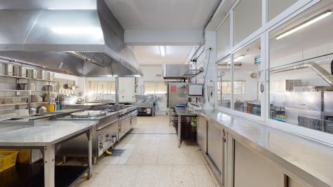Cocina. Cocina industrial amplia y muy luminosa, equipada por completo con mobiliario, encimeras y electrodomésticos de acero inoxidable. A la izquierda destacan los fogones tapados por una gran campana extractora metálica. El suelo es de baldosa clara y a la derecha hay un gran ventanal acristalado que comunica con otra estancia.