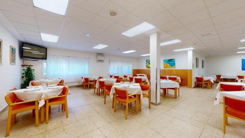 Comedor. Comedor amplio y muy luminoso con techo técnico blanco y suelo de baldosas beige. La sala está distribuida con múltiples mesas cuadradas vestidas con manteles blancos y rodeadas por sillas de madera con tapicería de color naranja vivo. Al fondo hay grandes ventanales con cortinas blancas, paredes decoradas con cuadros coloridos y una televisión colgada a la izquierda.