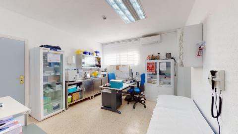 Consulta de enfermería. Despacho médico luminoso con paredes blancas y suelo de terrazo claro. En el centro destaca un escritorio gris con silla de oficina azul y un ordenador. A la izquierda hay una camilla de exploración con sábana blanca y un gran armario acristalado para medicamentos. Al fondo, una ventana con estor blanco ilumina la sala, equipada con armarios y material sanitario.