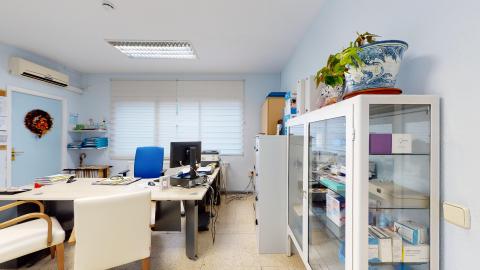 Consulta médica. espacho médico de paredes azul claro y suelo de terrazo. Cuenta con una mesa de escritorio de madera clara dispuesta en ángulo, con un ordenador, papeles y una silla de oficina azul. En primer plano a la derecha destaca un armario vitrina blanco con estantes de cristal llenos de cajas de medicamentos, coronado por una maceta de cerámica azul y blanca con una planta verde.