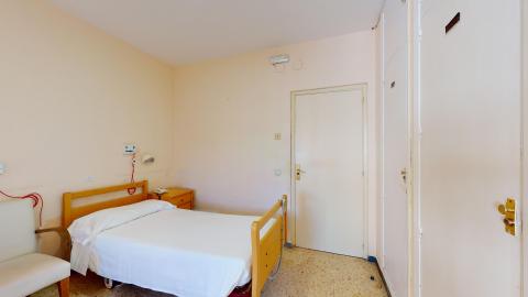 Habitación doble. Dormitorio de tonos beige y suelo de terrazo claro. Se muestra una cama articulada con cabecero de madera, sábanas blancas y un sistema de aviso de emergencia con cordón rojo en la pared. Al lado hay una mesilla de noche de madera y un sillón blanco. A la derecha se aprecian armarios empotrados blancos y la puerta de entrada.