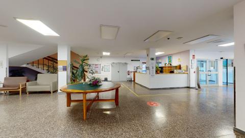 Recepción. Entrada principal espaciosa con suelo de terrazo marrón brillante y techos blancos con amplios paneles de luces fluorescentes. En el centro destaca una gran mesa ovalada de madera con un centro de flores. Al fondo a la derecha, se observa el mostrador blanco de la recepción y la puerta acristalada de entrada. A la izquierda, hay una escalera y varios sillones tapizados.
