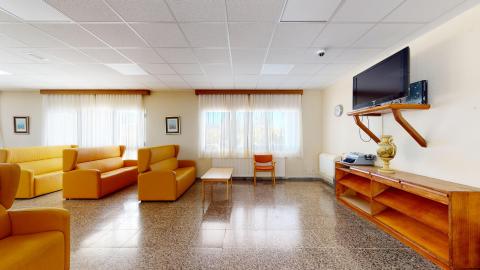 Sala de estar. Sala de estar muy amplia y luminosa, con paredes de color crema, techo técnico blanco y suelo de terrazo marrón reflectante. En la parte izquierda destacan varios sofás de tres plazas tapizados en un llamativo color amarillo mostaza. Al fondo, dos grandes ventanas con cortinas blancas dejan pasar la luz natural. A la derecha, un mueble bajo de madera soporta un televisor colgado en la pared.