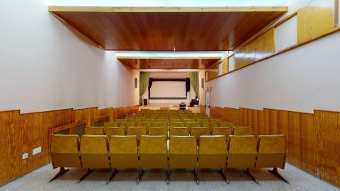 Salón de actos. Salón de actos o sala de proyecciones alargada, con paredes blancas y paneles inferiores de madera clara. Al fondo destaca un escenario con cortinas verdes y una gran pantalla de proyección en blanco. La sala está iluminada por una luz cenital indirecta que resalta las filas de butacas tapizadas en color mostaza, dispuestas en hileras orientadas hacia el escenario.