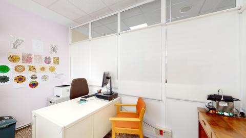 Consulta de psicología. Despacho luminoso con una pared lila claro decorada con numerosos dibujos de mandalas coloreados. Consta de una mesa de escritorio blanca con un ordenador de torre, una silla de oficina marrón y una silla confidente de madera con tapizado naranja. La pared del fondo está formada por paneles acristalados con estores blancos completamente bajados. Destaca una cafetera de cápsulas sobre un mueble auxiliar a la derecha.