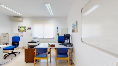 Trabajo social. Despacho de trabajo social con paredes blancas, suelo de baldosas claras y buena iluminación por fluorescentes y una ventana central con estor blanco. En el centro destaca una mesa de escritorio gris con dos sillas confidente tapizadas en azul brillante y una silla de oficina a juego. A la derecha resalta una gran pizarra blanca colgada en la pared y, al fondo, archivadores metálicos grises.