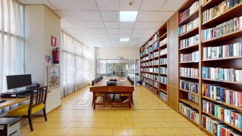 Biblioteca. Sala rectangular alargada y muy luminosa gracias a un gran ventanal con cortinas blancas a la izquierda. El suelo es de baldosas amarillas y las paredes son de tono crema. A la derecha destaca una gran estantería de madera repleta de libros ordenados por letras. En el centro, una mesa alargada de madera con sillas y, en primer término a la izquierda, un puesto con ordenador de mesa.