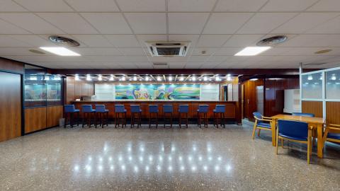 Cafetería. Sala amplia con suelo de terrazo brillante de tonos grises y marrones. Al fondo destaca una larga barra de cafetería de madera oscura iluminada por una hilera de focos empotrados y decorada con un gran mural abstracto de colores vivos (azules, verdes y amarillos). Frente a la barra y a la derecha hay taburetes y sillas tapizadas en un llamativo color azul. La iluminación del techo es moderada.